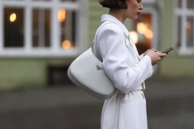 A person in a white coat using a smartphone with a white handbag on shoulder, standing outdoors.