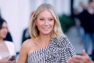 A woman with blonde hair in a striped, ruffled, off-shoulder dress at an event, smiling while looking away from the camera.