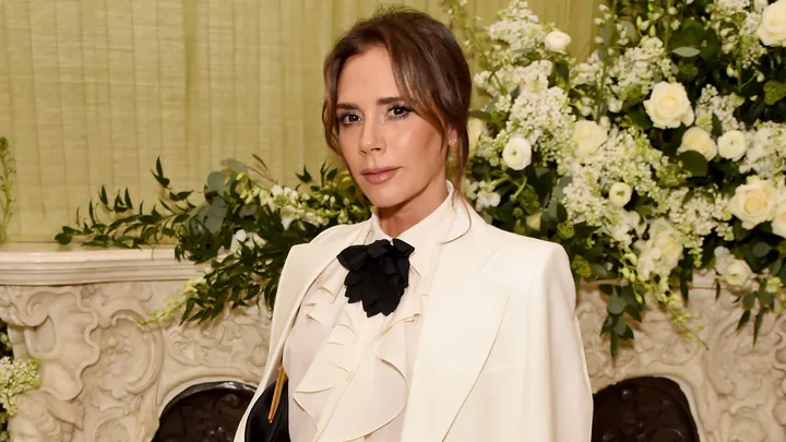 A woman in a white suit with a black bow stands in front of a floral arrangement indoors.