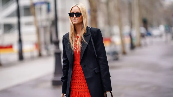 Woman in sunglasses wearing a black jacket over a red dress, walking down a city street.