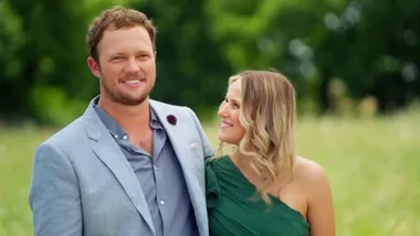 A man wearing a light blue suit and a woman wearing a green dress smiling at each other in a field.