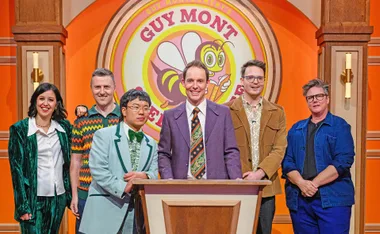Guy Montgomery, in a purple jacket and a tie, stands behind a podium, with his sidekick Aaron Chen and competitors behind him.